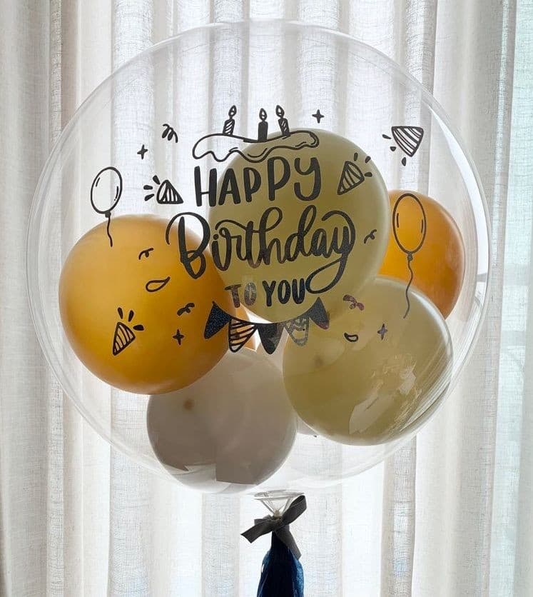 Personalized clear bubble balloon reading HAPPY BIRTHDAY TO YOU, stuffed with gold and white balloons, floating against a white curtain