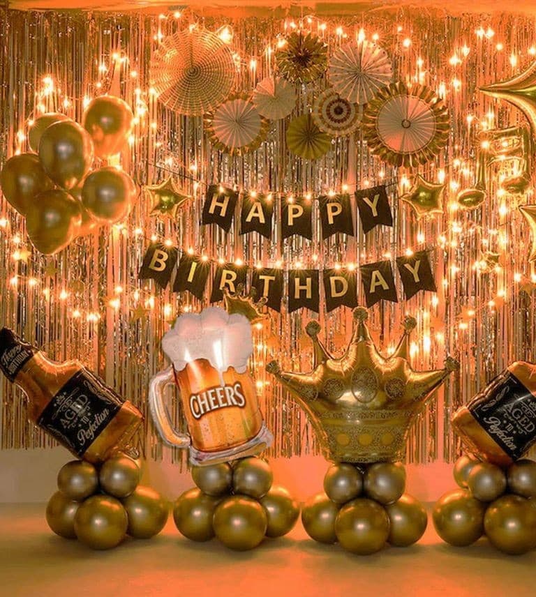 Gold and black birthday setup: a HAPPY BIRTHDAY banner over a gold fringe curtain, with crown and drinks-themed mylar balloons and gold balloon clusters on the floor