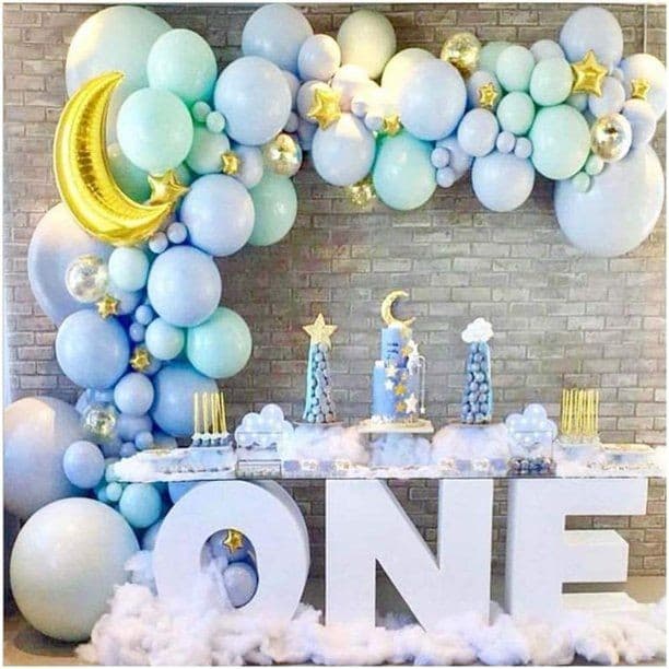 First birthday setup: a pastel blue, mint and gold organic balloon garland over a brick wall, with a large ONE letter display, gold moon and a cloud-styled cake table