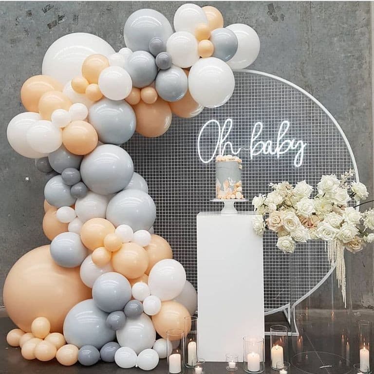 Baby shower backdrop: an organic balloon garland in peach, grey and white curving around a round mesh arch with a neon "Oh baby" sign, white roses, a cake pedestal and candles on the floor