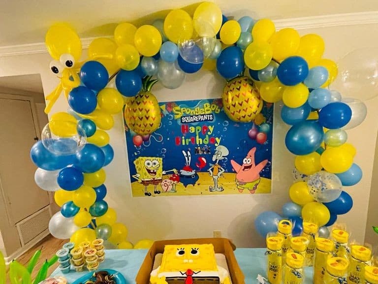 SpongeBob themed birthday arch in yellow and blue with pineapple foil balloons and character eyes, framing a SpongeBob backdrop above a themed cake table