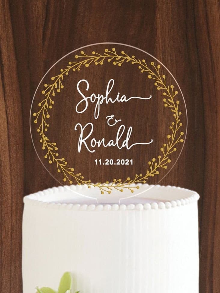 Clear acrylic cake topper engraved with "Sophia & Ronald, 11.20.2021" inside a gold laurel wreath, set on a white wedding cake